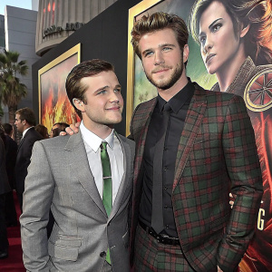 Josh Hutcherson and Liam Hemsworth at a film premiere. Josh in a gray suit with green tie, Liam in a plaid jacket over black shirt. Friendly pose, vibrant movie poster background.