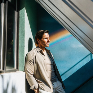 Sam Jaeger, actor, posing against a vibrant rainbow backdrop. He is dressed in a light brown shirt with neatly combed short hair.