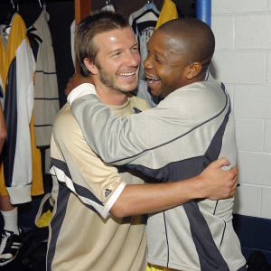 David Beckham and Albert Morgan, dressed in soccer jerseys, in a locker room filled with team gear. They are laughing and hugging. Warm, friendly atmosphere