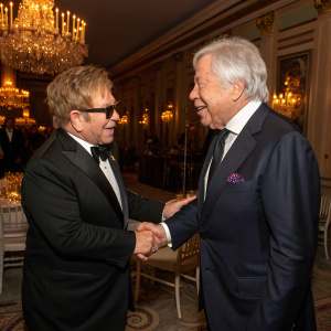 Sir Elton John and Robert Kraft shaking hands at a private gala. Lavish interior with chandeliers