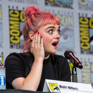 Maisie Williams, with vibrant pink hair in a high ponytail, looking astonished at a bustling Comic-Con event. Dressed in a black top and adorned with red nail polish, she stands before a backdrop filled with Comic-Con branding. Candid, close-up shot.