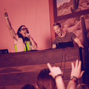 Skrillex and Diplo performing at Coachella. Skrillex in neon outfit, Diplo in casual wear. Desert landscape backdrop