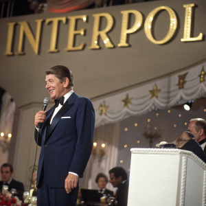 Ronald Reagan, in a navy suit, addresses an audience at a gala. Behind him is a stage set with golden letters spelling out 'Interpol'.
