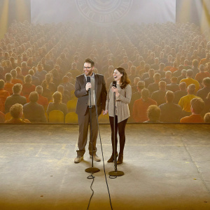 Seth Rogen and Lauren Miller Rogen, hosting a comedy fundraiser. Crowded auditorium, spotlighted stage.