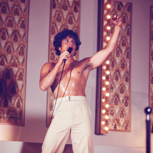 Bruno Mars, shirtless, performing on stage. Muscular build, curly dark hair. Vibrant concert backdrop.