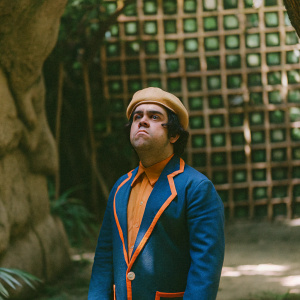 Don Ramón from El Chavo del Ocho, stern expression, wearing a vibrant blue and orange outfit. Weird patterned backdrop.