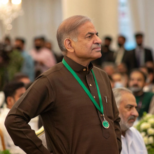 Close-up of Shehbaz Sharif in brown shirt with green lanyard. Formal event. Crowd in background blurred