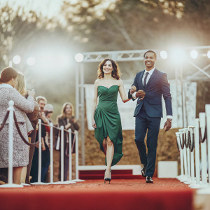 Simon Webbe and Ayshen Kemal at a glamorous awards show. Simon in a sharp navy suit, Ayshen in a stunning emerald green dress. Bright stage lights, audience in the background.