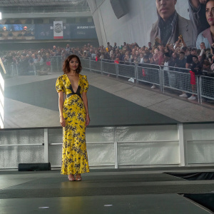 Zendaya at New York Comic Con, dressed in a vibrant yellow floral print dress, standing in front of a crowd of fans and media. Event backdrop