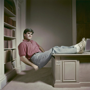 Sean Hannity, Fox News host, appears in a casual setting. Jeans, red polo shirt. Office desk in the background, bookshelves and laptop visible