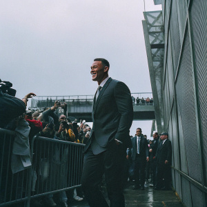 Aaron Judge, in a sleek black suit, at a post-game press conference. Cheerful crowd in background.