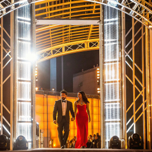 Frank Grillo and a woman with dark, sleek hair at an awards ceremony. Both dressed in glamorous evening wear. Bright stage lights and audience in the background.