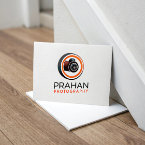 A modern logo for a photography business named 'Prahan Photography'. It features a sleek camera lens icon centered on a minimalist white background with an accent of bold orange ring. The business name is displayed below in a stylish font.