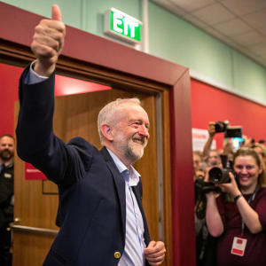 Jeremy Corbyn, sporting a dark suit and cheerful smile, giving a thumbs up. The background is filled with an excited crowd capturing the moment with cameras and phones. The scene is set in a vibrant indoor event with a wooden door and green exit sign.