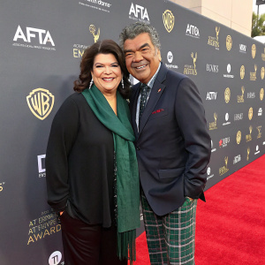 Ann Lopez and George Lopez at award show. Ann in dark suit and green scarf. George in collared shirt and plaid pants. Both smiling and posing on red carpet surrounded by sponsors' logos.