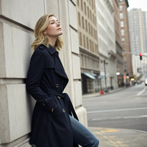 Rosamund Pike in a candid photo shoot. Wearing a black trench coat and jeans. Grey urban background.