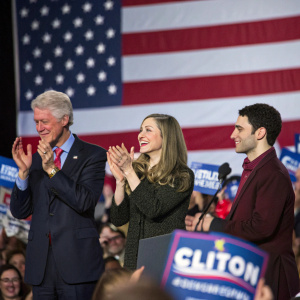 Bill Clinton, Chelsea Clinton, and Marc Mezvinsky clapping at a political rally. American flag waving in the background. Crowded and enthusiastic atmosphere
