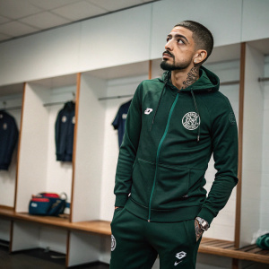 Manuel Lanzini, midfielder, standing in the locker room. Dark green tracksuit with QPR logo. Intense expression. Tattoos visible on his forearms