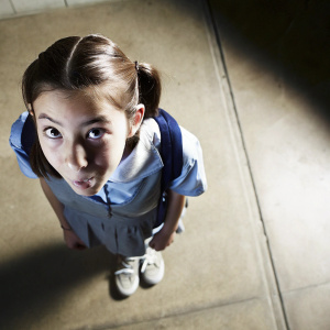 High-angle shot of a young Asian girl with playful expression. Pigtails, school uniform, casual shoes. Plain background, close-up of upper body and face.