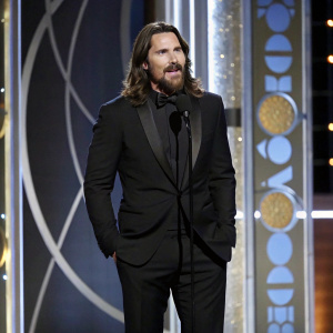 Christian Bale, in a tuxedo, with a scruffy beard and long hair, addressing the audience on the Golden Globe Awards stage.