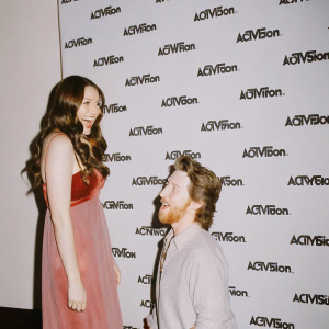 Clare Grant and Seth Green at the Activision event. Clare with long brown hair, cheerful smile, dressed in a red carpet gown. Seth in casual attire with beard. Black backdrop with repeating Activision logos.