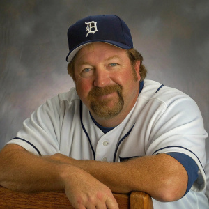 Professional photo of Alan Zinter. Caucasian, middle-aged, full beard and mustache. Wearing a white baseball uniform with navy blue accents and a matching navy blue baseball cap featuring a team logo. Relaxed and friendly demeanor. Shot against a subtle gray background.