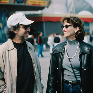 Steven Spielberg and Kate Capshaw at E.T. Adventure opening event. Spielberg in tan jacket, dark t-shirt, white cap. Capshaw in black leather jacket, pearl necklace, sunglasses. Vibrant backdrop