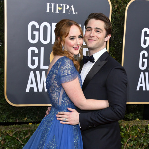 Camryn Grimes and Oliver Singer locked in an embrace at the Golden Globe Awards. Formal attire, black tuxedo, blue gown with lace detailing. Dark, glamorous backdrop with award logos.