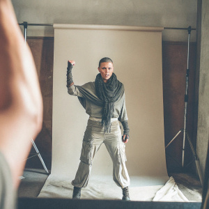 Imperator Furiosa from Mad Max: Fury Road, fierce look. Grayish outfit with dark scarf. Shot in a studio with gray backdrop.