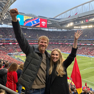 Alexi Lalas and Maria Komandnaya at a bustling stadium, cheering for their team amidst a crowd of enthusiastic fans.