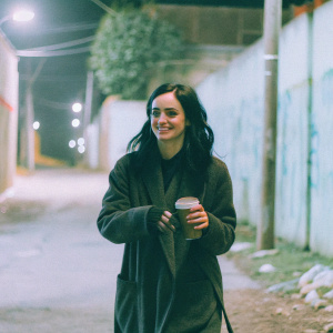 Krysten Ritter, walking casually through a dimly lit alleyway, wearing a dark overcoat, with a friendly smile. The background is blurred, showing glimpses of graffiti and street lamps.