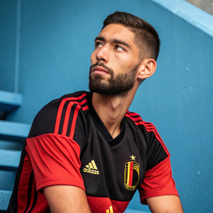 Professional soccer player Yannick Ferreira Carrasco poses formally. Wearing black and red jersey with Adidas branding. Serious expression. Solid blue background.