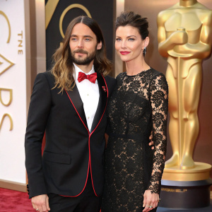 Jared Leto in a Gucci suit, red bow tie, standing next to a woman in a sophisticated lace dress, both posing formally. Male actor in sleek black tuxedo, crisp white shirt. Oscar statuette on display. Bright red carpet background, high resolution.
