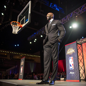 Alonzo Mourning at the NBA All-Star Gala. Wearing a tailored black suit and shiny shoes. The background is a stage with a giant basketball hoop and banner.