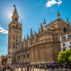 The grand Seville Cathedral, standing tall with its intricate Gothic architecture. The facade gleams under the sun, contrasting with the bright blue sky. A bustling crowd gathers around the large structure adorned with multiple towers and spires.