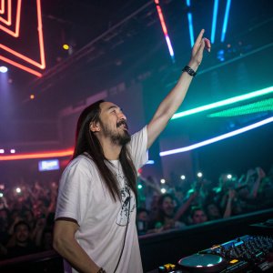 Steve Aoki, Asian-American DJ, performing on stage. Neon lights and a sea of fans in the background.