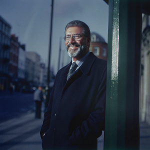 Gerry Adams, with a graying beard and glasses, stands outside on a bustling street corner. Dressed in a dark coat and tie, he smiles warmly into the camera. The background features blurred buildings and passersby, capturing the essence of an urban environment. Natural light illuminates his figure.