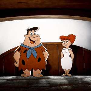 Cartoon illustration featuring Fred Flintstone, big and smiling, dressed in brown stone-like attire with a blue scarf. Beside him stands Wilma Flintstone, curvy and elegant, clad in a simple white outfit. Traditional animation style with a plain white backdrop.