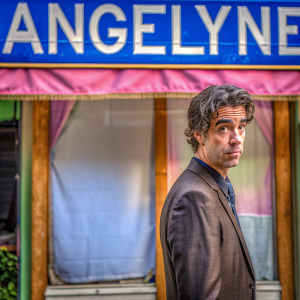 Hamish Linklater, Caucasian actor, in a dark suit with a frown. Pink fabric backdrop with large 'ANGELYNE' text. Film festival event.