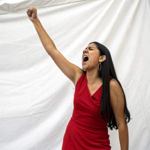Illustration of a Hispanic woman in her 20s, with long straight black hair, wearing a red sleeveless dress. She is depicted with a raised arm and a loud, expressive yell against a plain white backdrop.