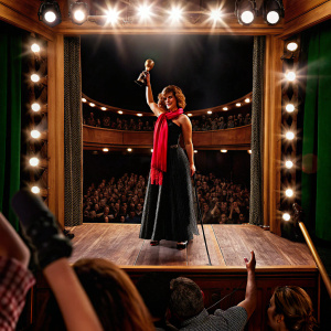 Shania Twain accepting a Grammy award. Wearing black dress, red scarf. Crowded auditorium with flashing lights