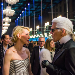 Naomi Watts and Karl Lagerfeld at a glitzy gala. They are engaged in animated conversation. The setting is dimly lit with chandeliers. Background filled with socialites and celebrities.
