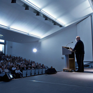 Economist Joseph Stiglitz delivering a keynote speech at a university conference. Brightly lit auditorium with audience in attendance