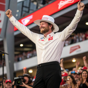 Dale Earnhardt Jr., smiling broadly in victory lane. Wearing a sleek white shirt with red logos, black pants, and a matching hat. Crowds of cheering fans in the background.