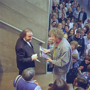 James Lipton and Gene Wilder at a book signing event. Lipton with a gray beard, wearing a black suit. Wilder in a casual gray jacket. They are surrounded by eager fans and signed copies of their books.