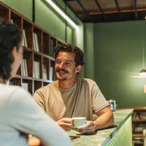 Orlando Bloom, wearing a casual, beige t-shirt, hanging out in a cozy coffee shop. Warm lighting and bookshelves in the background.