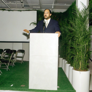 Ralph de la Vega, bearded man in navy suit, addressing an audience at a tech conference. Sparse stage design
