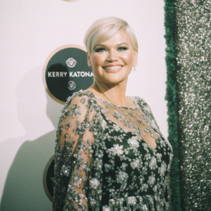 Kerry Katona, with a radiant smile and short blonde hair, attending the glamorous awards ceremony. Wearing a stunning floral-patterned dress adorned with crystals. The backdrop features a striking mix of purple and white hues, interspersed with logos and event text.
