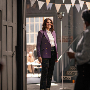Megan Mullally, dressed in a purple blazer, white shirt, and patterned black pants, attending the Tribeca TV Festival. Festive backdrop filled with event branding.