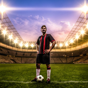 Theo Hernandez, soccer player, poses in AC Milan's jersey. Black with red accents, standing on a soccer field. Stadium lights, blurred crowd in background.